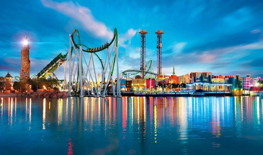 Panorâmica noturna do parque Islands of Adventure, com a montanha-russa The Incredible Hulk iluminada e refletida nas águas do lago central.