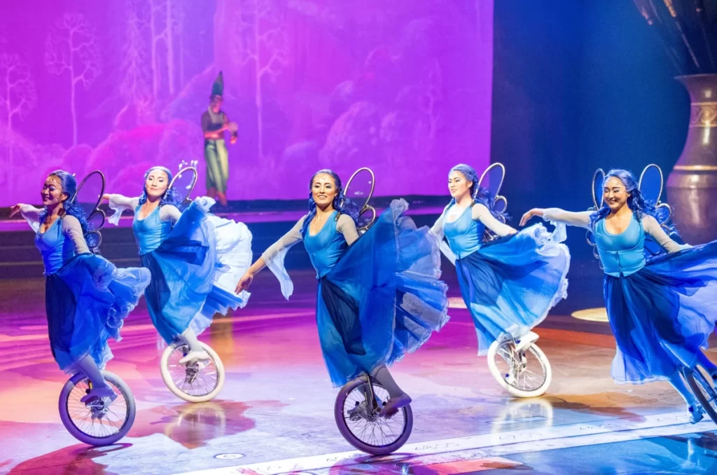 Artistas em monociclos vestidos de fadas azuis se apresentam no palco com projeções ao fundo.