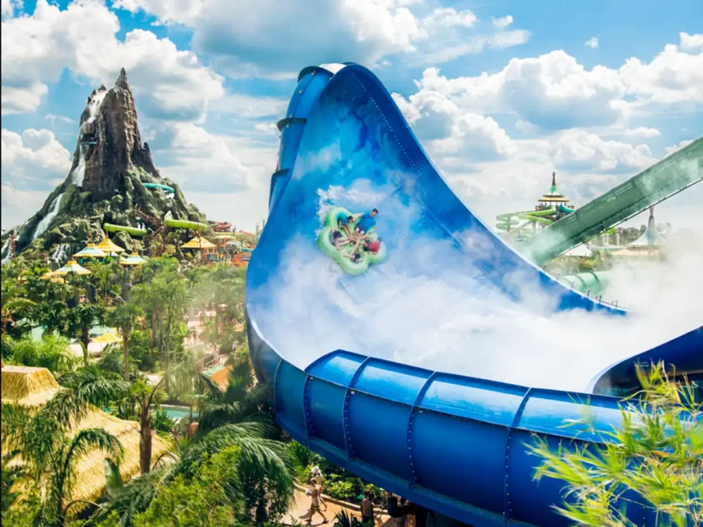 Visitantes descem um toboágua azul com o vulcão ao fundo no Volcano Bay.