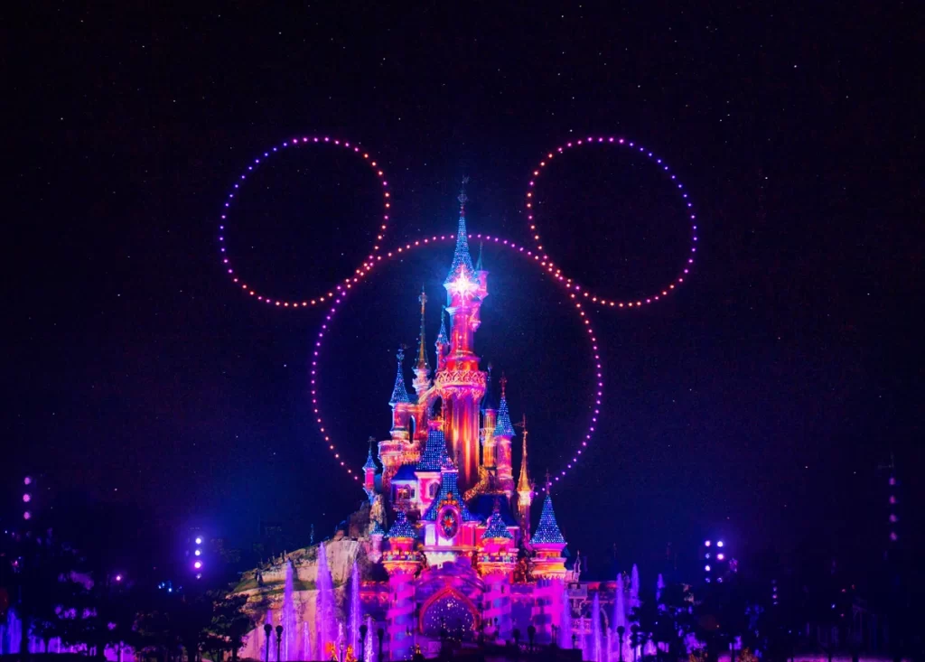 Castelo da Disneyland Paris iluminado com fontes dançantes e drones desenhando a silhueta do Mickey ao fundo.