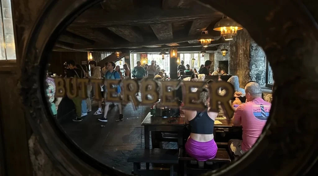 Interior do Cabeça de Javali visto através de janela com inscrição “Butterbeer”, mostrando clientes e balcão do pub.