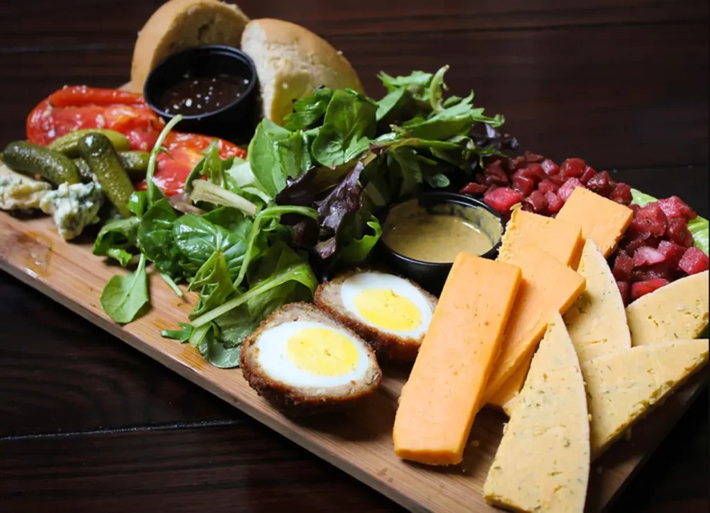 Prato Ploughman’s do Leaky Cauldron com queijos, ovos empanados, salada, picles, pães e chutney, servido em tábua de madeira.