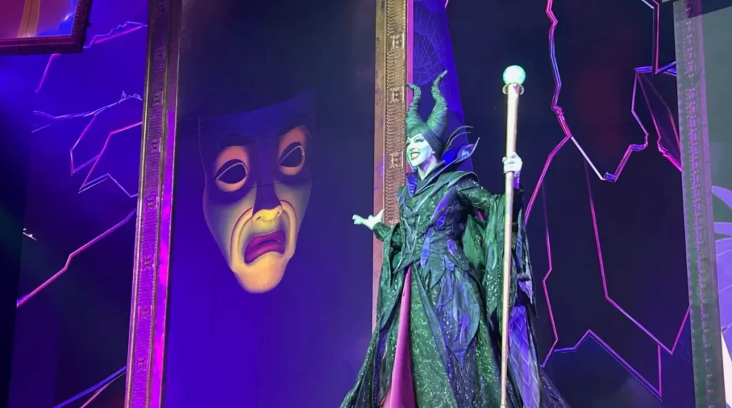 Malévola sorrindo ao lado da máscara falante gigante, iluminação roxa e atmosfera teatral.