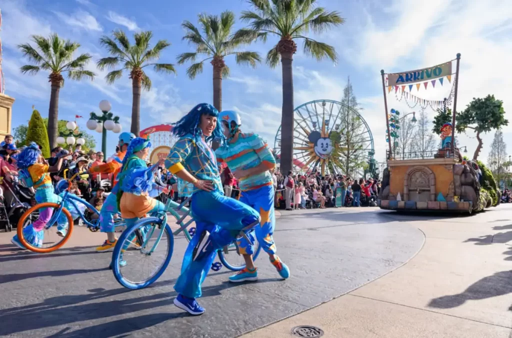 Dançarinos e ciclistas coloridos animam o desfile Pixar Pier na Disney California Adventure sob céu ensolarado.