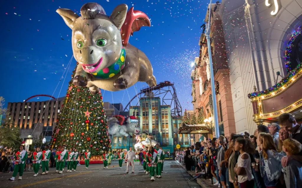 Balão gigante de personagem no desfile de Natal do Universal Orlando, com decoração luminosa e público animado.