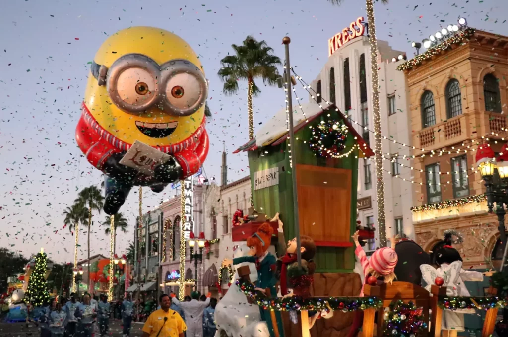 natal epic universe - Balão dos Minions no desfile de Natal do Universal Studios Florida com confetes e luzes festivas na rua principal.
