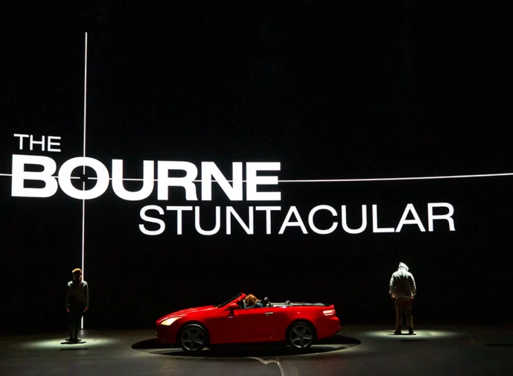 Palco da atração The Bourne Stuntacular com carro vermelho e iluminação dramática.