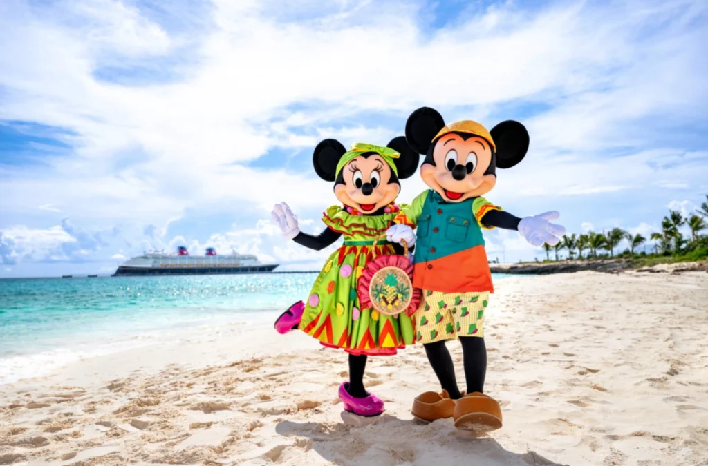 Mickey e Minnie com roupas tropicais posando na praia de Disney Lookout Cay com navio ao fundo.