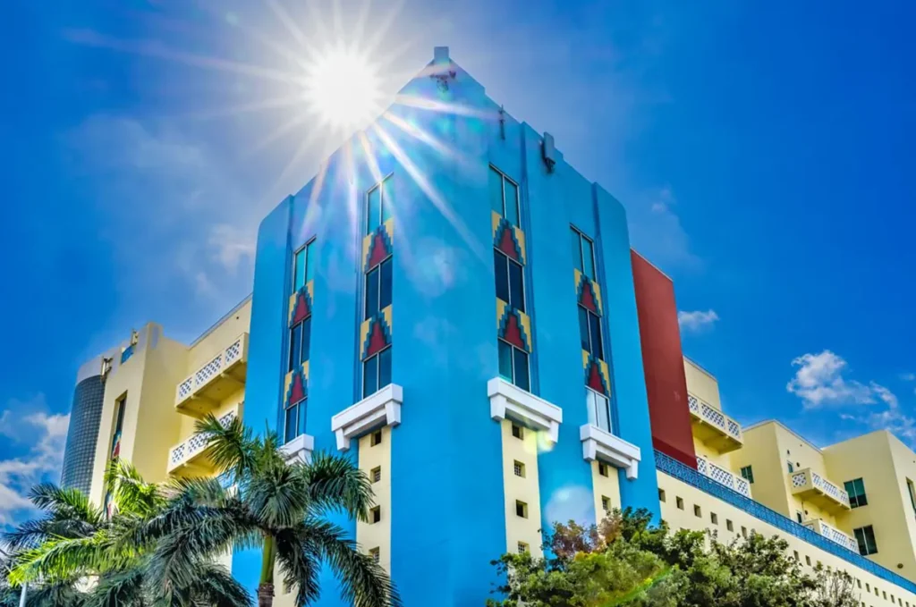 Fachada azul de hotel Art Déco iluminada pelo sol, com palmeiras e detalhes geométricos característicos.