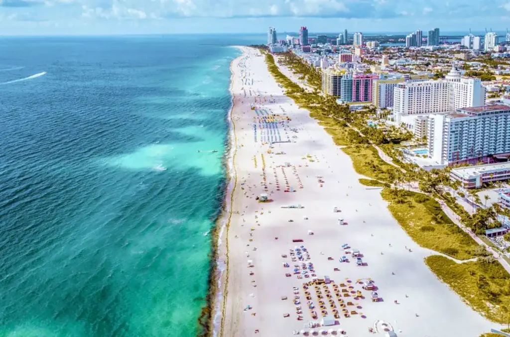 Litoral de Miami Beach visto do alto, com praia de areia branca, mar azul-turquesa e hotéis ao longo da costa.