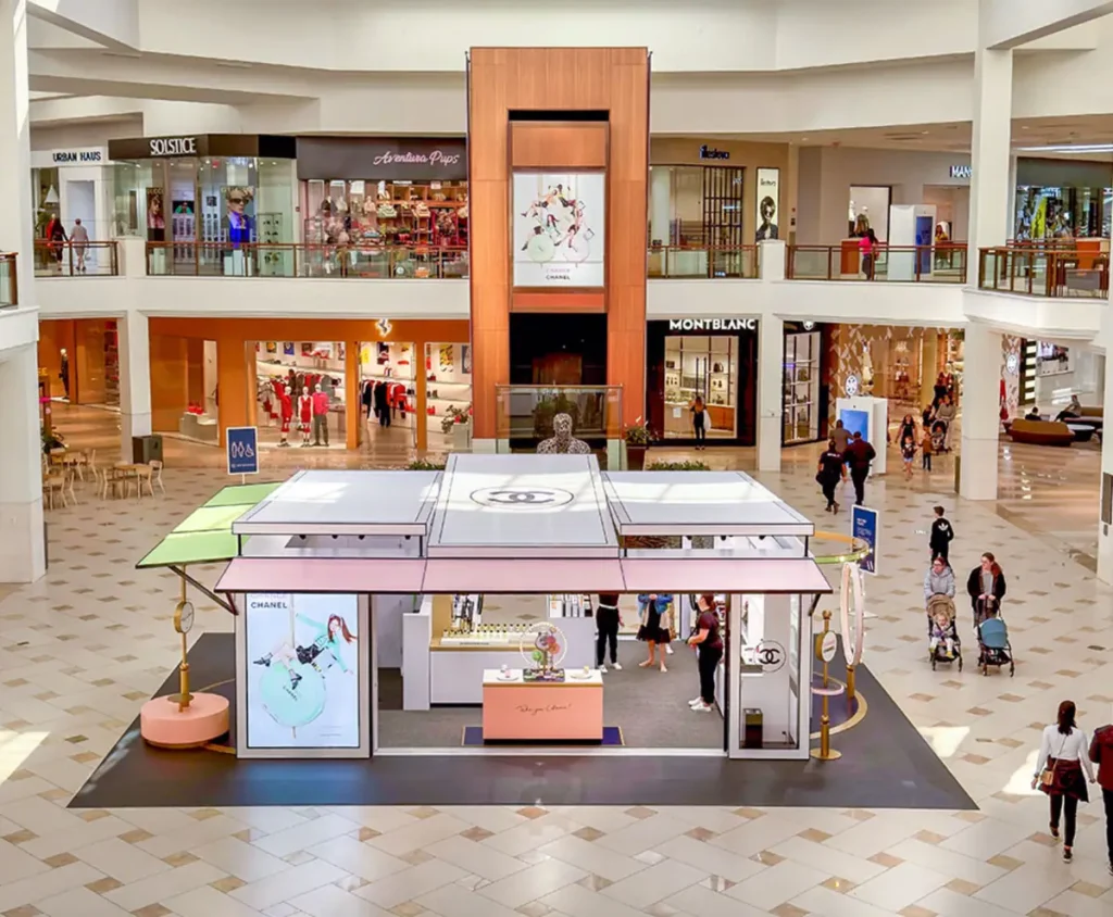 Interior do Aventura Mall com dois andares de lojas de luxo, visitantes circulando e um quiosque da Chanel no centro do átrio.