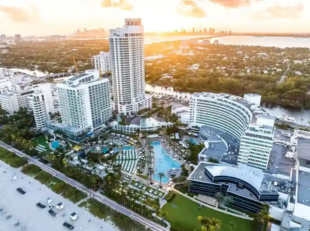 Fontainebleau Miami Beach visto do alto ao pôr do sol, com piscinas, praia e skyline de Miami ao fundo.