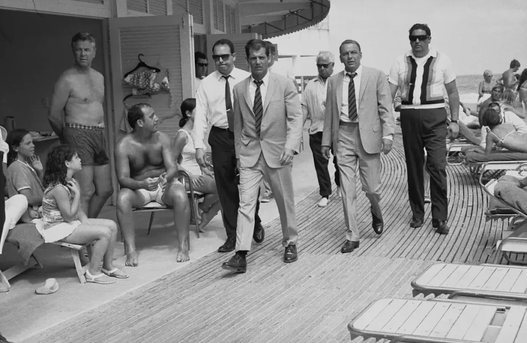 Frank Sinatra, ao centro e à frente da imagem, caminha com sua comitiva pela área da piscina do Fontainebleau Miami Beach durante as filmagens de The Lady in Cement, em 1968. A fotografia é de Terry O’Neill