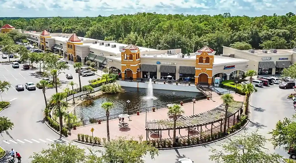 Vista panorâmica do Lake Buena Vista Factory Stores, em Orlando, destacando suas lojas, estacionamento e área de convivência com lago e fonte.